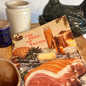 May include: A vintage cookbook titled The Best of Bacon featuring a large raw pork roast in the foreground, a pineapple, a copper pot, and a butter stick. The cover art displays a culinary scene with various kitchen items and food ingredients.