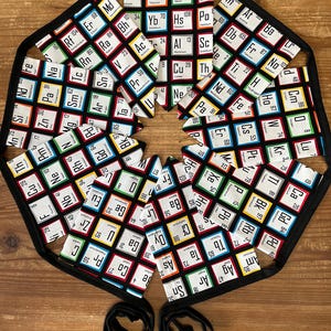 May include: An octagonal black apron with a periodic table design. The apron has a black border and two black ties. The periodic table elements are displayed in colourful squares on a white background.