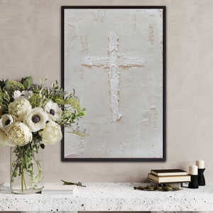 Puede incluir: Una cruz blanca está pintada sobre un lienzo blanco texturizado, enmarcado en negro. El lienzo está colgado en una pared beige claro sobre una mesa blanca con un jarrón de flores blancas y libros.