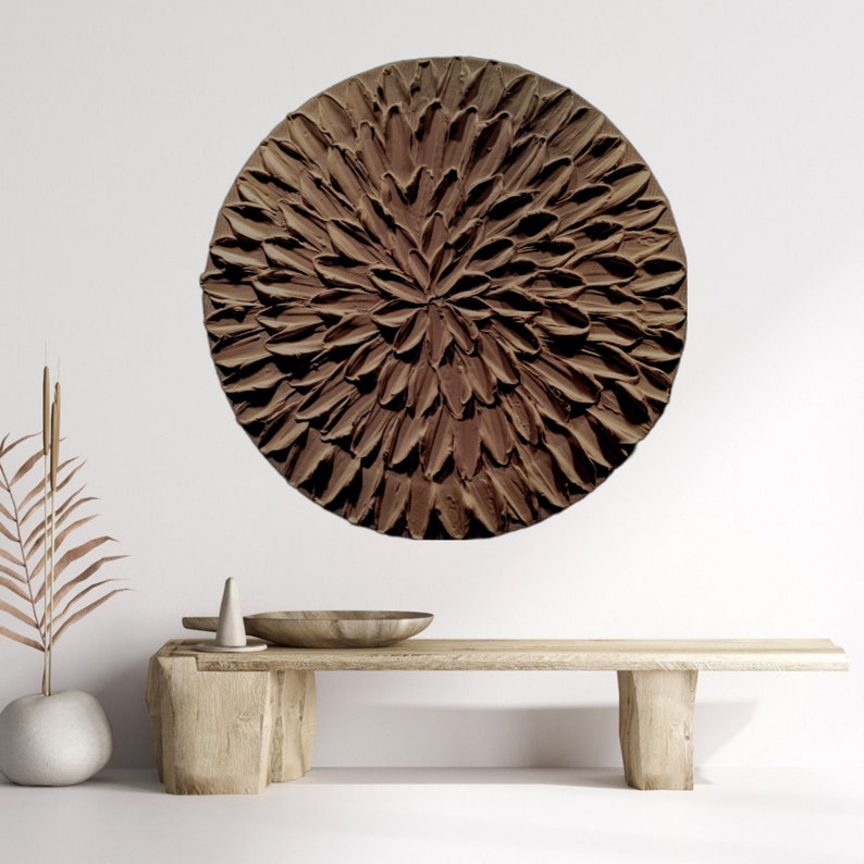 May include: A large, round, brown wall art piece with a textured, flower-like design. The art piece is hanging on a white wall above a wooden bench.