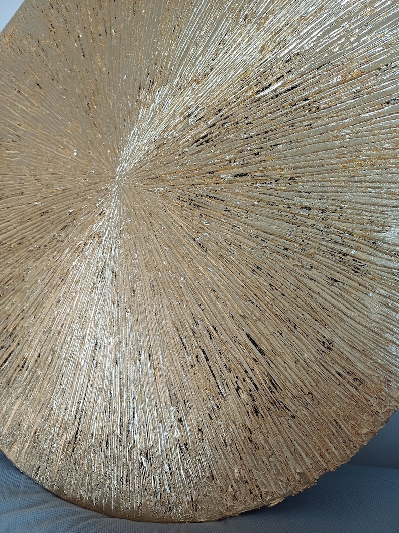 May include: A round, gold-colored wall decor with a textured, sunburst design. The surface is covered in raised lines that radiate outward from the center.