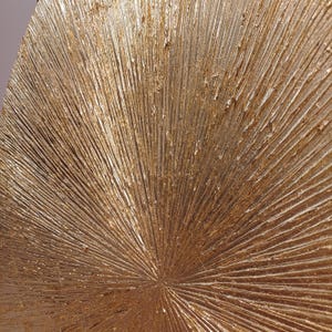 May include: A gold-toned, textured, circular wall decor with a radiating pattern. The surface has a rough, textured look, resembling a sunburst.