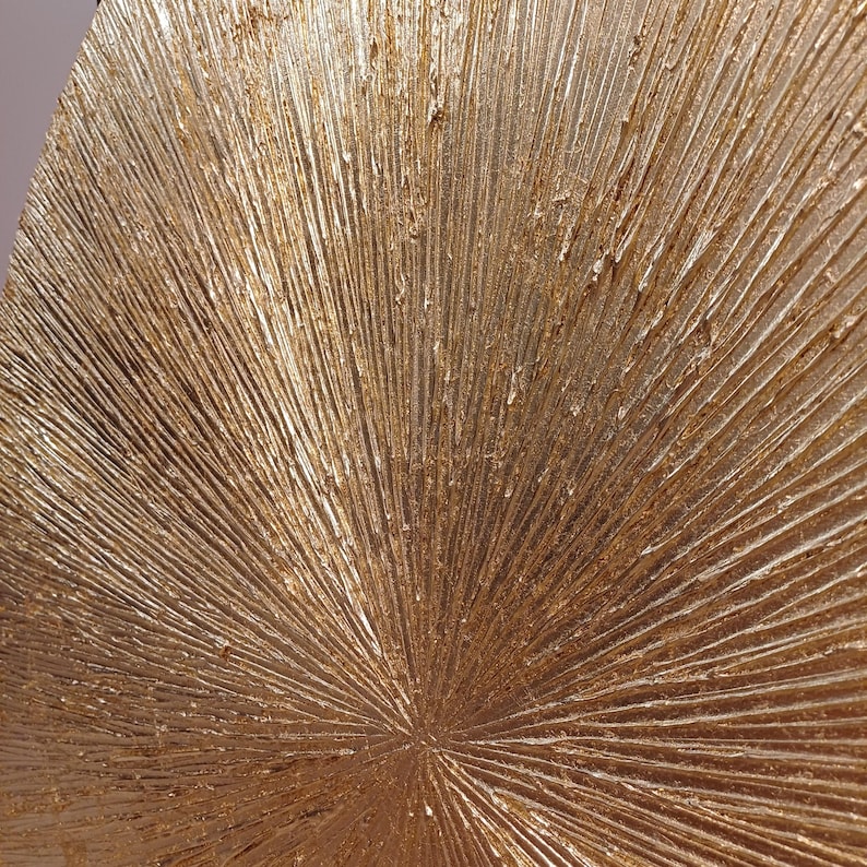 May include: A gold-toned, textured, circular wall decor with a radiating pattern. The surface has a rough, textured look, resembling a sunburst.