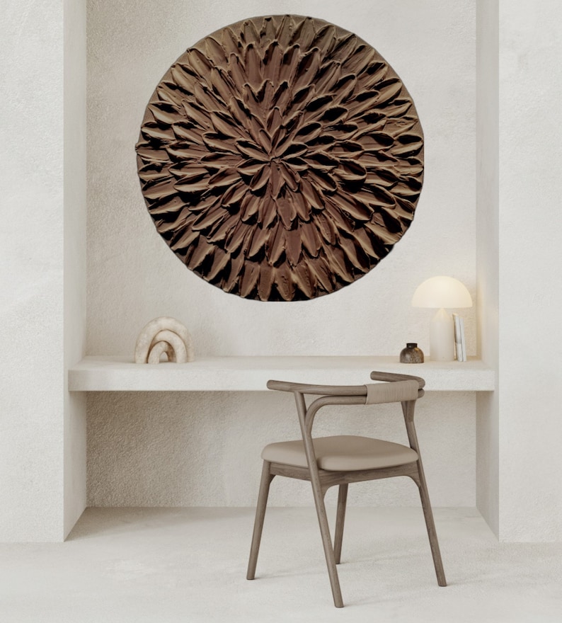 May include: A brown circular wall art piece with a textured, flower-like design hangs above a white desk with a wooden chair.