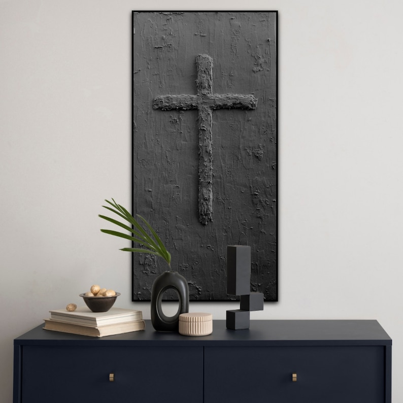 Peut inclure: Une peinture abstraite en noir et blanc d'une croix sur un fond textur&eacute;. La peinture est encadr&eacute;e d'un cadre noir et est accroch&eacute;e &agrave; un mur blanc. La peinture est au-dessus d'une commode bleu fonc&eacute; &agrave; deux tiroirs. La commode est d&eacute;cor&eacute;e d'un vase, d'un bol de noix et d'un livre.