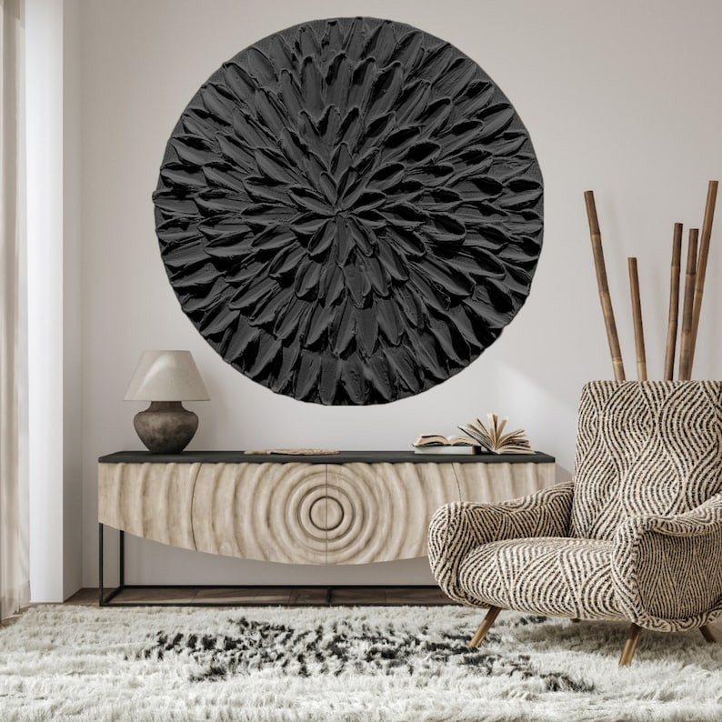 May include: A black and white abstract wall art piece with a circular shape and a textured, 3D design. The art piece is hanging on a white wall above a wooden console table with a black top and a patterned front.