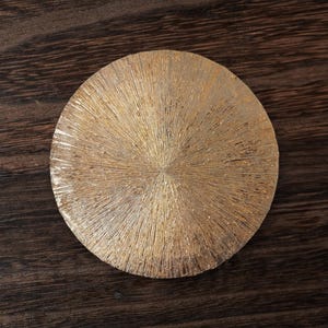 May include: A round, gold-colored coaster with a textured surface. The coaster has a radial pattern that resembles a sunburst.