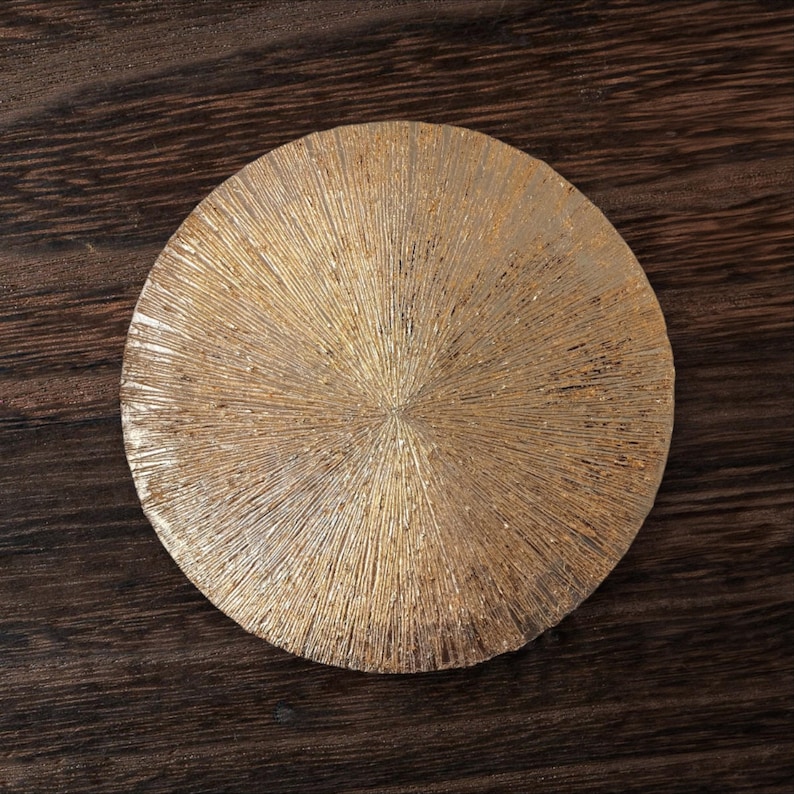May include: A round, gold-colored coaster with a textured surface. The coaster has a radial pattern that resembles a sunburst.
