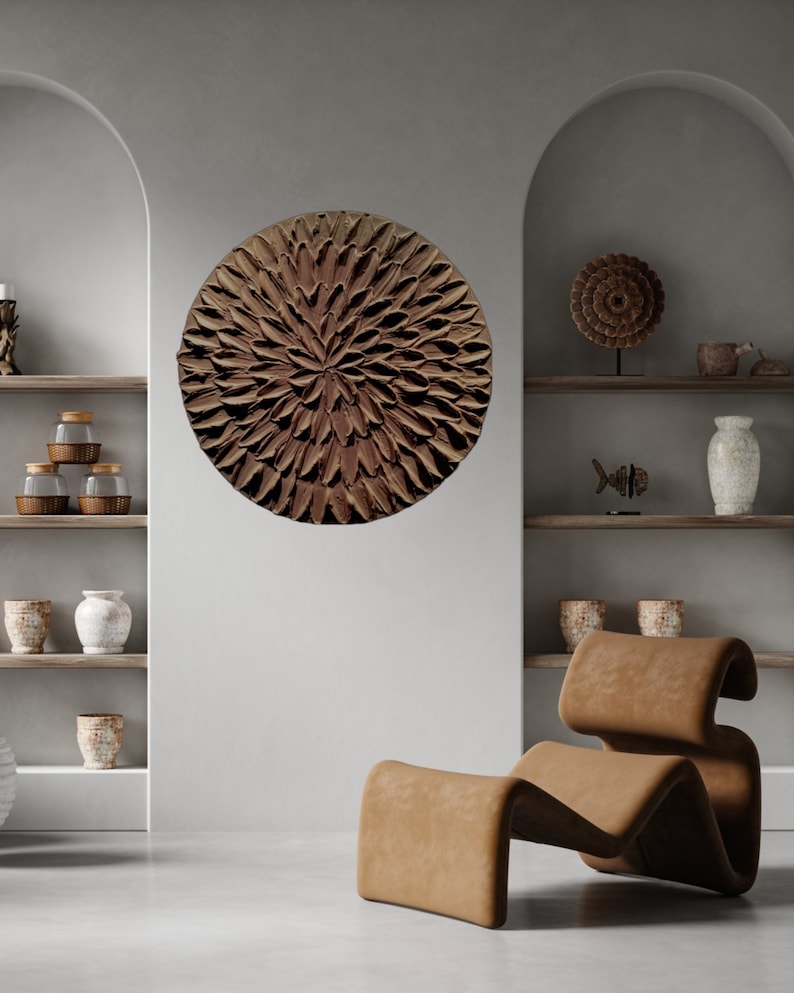 May include: A brown circular wall art piece with a textured, flower-like design. The art is hanging on a white wall in a room with a brown leather chair.