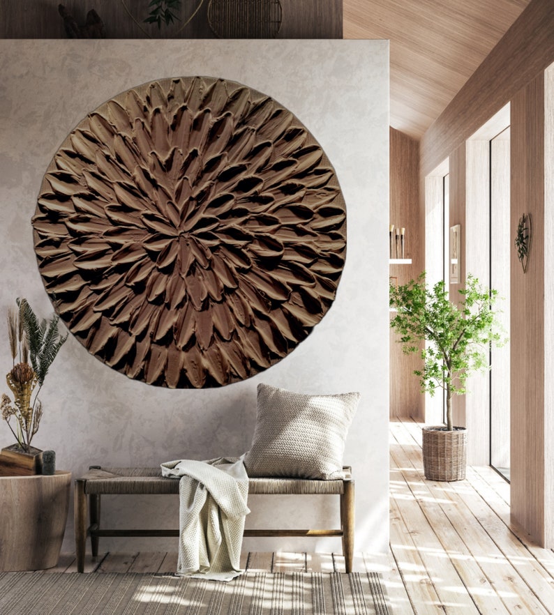 May include: A large, round, brown wall art piece with a textured, flower-like design. The art is hanging on a light gray wall in a modern living room.