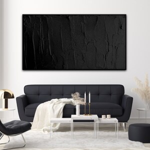 Puede incluir: Una pintura abstracta en blanco y negro con una superficie texturizada cuelga de una pared blanca sobre un sof&aacute; negro y mesas de caf&eacute; blancas.