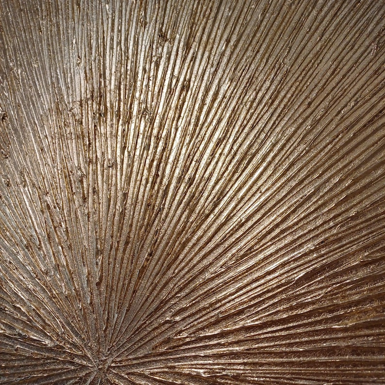 May include: A close-up of a textured, metallic gold surface with a radial pattern. The surface has a raised, brushstroke-like texture, creating a dimensional effect.