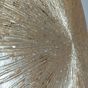 May include: A close-up of a round, textured, gold-colored wall art piece. The surface is covered in raised lines that radiate outward from the center.