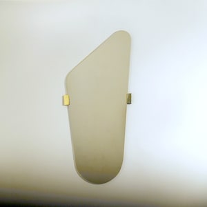 Può includere: Uno specchio grande, di forma irregolare, di colore bianco sporco, con un bordo inferiore arrotondato. Lo specchio è montato su una parete bianca con due staffe in metallo dorato.