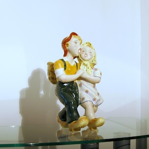 May include: A ceramic figurine of a boy and girl holding hands. The boy is wearing a yellow shirt, green trousers, and a red hat. The girl is wearing a white dress with yellow flowers and a yellow headscarf.