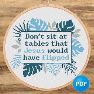 May include: A cross-stitch embroidery hoop with the text "Don't sit at tables that Jesus would have flipped." The design features blue and gray leaves and foliage around the text. The background is white, and the hoop is made of wood.