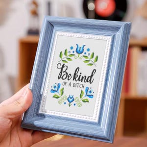 May include: A framed cross-stitch artwork with a light blue frame. The artwork features the text "Be kind of a bitch" in black script, surrounded by blue floral and green leaf accents. The frame is held by a hand.