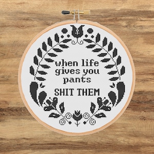 May include: A circular wooden embroidery hoop containing white fabric with black cross-stitch embroidery. The design features a floral wreath surrounding the text: when life gives you pants SHIT THEM. The embroidery is centred within the hoop.