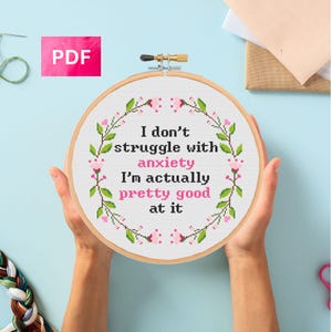 Pode incluir: Um bastidor de bordado em ponto cruz com o texto "I don't struggle with anxiety, I'm actually pretty good at it." O design apresenta uma coroa de flores em verde e rosa. Um ícone PDF rosa está no canto superior esquerdo.