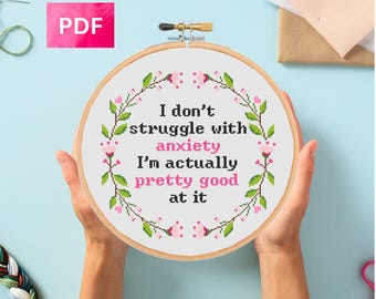 May include: A cross-stitch embroidery hoop with the text "I don't struggle with anxiety, I'm actually pretty good at it." The design features a floral wreath in green and pink. A pink PDF icon is in the upper left corner.