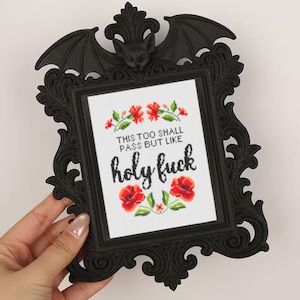 May include: Black framed cross-stitch art with a bat and floral design. The artwork features the phrase "THIS TOO SHALL PASS BUT LIKE holy fuck" in black text, surrounded by red flowers and green leaves.