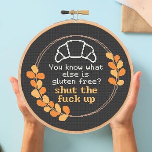 May include: A cross-stitch embroidery hoop with a black background and a white croissant graphic. The text reads, "You know what else is gluten free? shut the fuck up." Orange and yellow leaf accents surround the text.