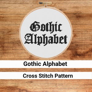 Puede incluir: Un aro de bordado blanco con letras de punto de cruz negro que dice "Gothic Alphabet" sobre un fondo de madera.