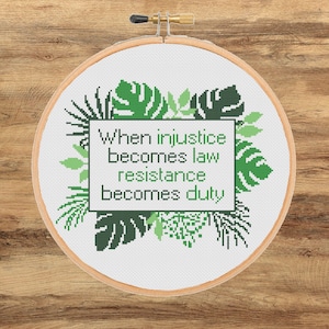 May include: A cross-stitch embroidery hoop featuring the quote "When injustice becomes law resistance becomes duty." The text is in a white rectangle, surrounded by green leaf designs. The hoop is set against a wooden background.