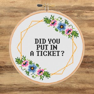 May include: A cross-stitch embroidery hoop with the text "DID YOU PUT IN A TICKET?". The design includes a gold geometric shape and floral accents with pink and blue flowers. The hoop is set against a wooden background.