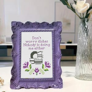 May include: A framed cross-stitch with a purple ornate frame. The cross-stitch reads "Don't worry dishes Nobody is doing me either" above a graphic of dishes and flowers. The frame is on a white surface.