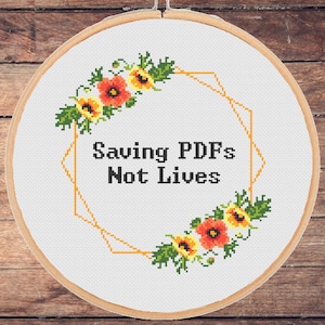 May include: A white embroidery hoop with a gold geometric frame and floral accents. The text "Saving PDFs Not Lives" is stitched in black thread.