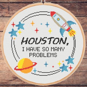 Op de afbeelding: Witte borduurring met een kleurrijke raket, sterren en planeten. De tekst luidt "Houston, ik heb zoveel problemen."