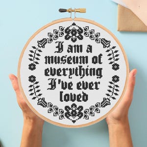 May include: A round, wooden embroidery hoop holds a completed cross-stitch piece. The text "I am a museum of everything I've ever loved" is stitched in black on white fabric, surrounded by floral and geometric patterns.
