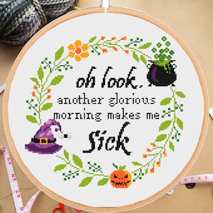 May include: A white cross-stitch embroidery hoop with a Halloween-themed design. The design features a witch's hat, a cauldron, a pumpkin, and the text "Oh look, another glorious morning makes me sick."