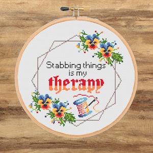May include: A circular wooden embroidery hoop containing white fabric with a cross-stitch design. The text reads Stabbing things is my therapy in black and red letters, surrounded by colorful pansy flowers and a spool of thread with a needle.