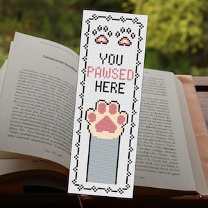 Op de afbeelding: Een witte bladwijzer met een zwarte rand en een kattenpootontwerp. De tekst "YOU PAWSED HERE" is roze. De bladwijzer is in een open boek geplaatst.