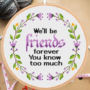 Może przedstawiać: Obręcz do haftu krzyżykowego w kolorze fioletowym i zielonym z tekstem "We'll be friends forever You know too much". Obręcz jest otoczona wieńcem kwiatowym z fioletowymi kwiatami i zielonymi liśćmi.