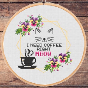 Op de afbeelding: Borduurpatroon met een kattengezicht, een kopje koffie en de tekst "Ik heb nu koffie nodig miauw" omringd door een gouden geometrisch frame en paarse en gele bloemen.