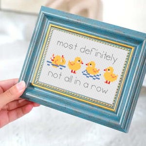 Puede incluir: Una obra de arte de punto de cruz enmarcada con un marco de madera azul. La obra presenta cuatro patitos amarillos en el agua, con el texto "most definitely not all in a row". El marco tiene un borde interior dorado.
