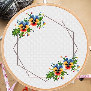 May include: A cross-stitch pattern featuring a geometric frame with two clusters of yellow, blue, and red pansies on a white background.