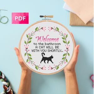 May include: A cross-stitch hoop with the text "Welcome to the bathroom A cat will be with you shortly..." surrounded by a floral design. A black cat silhouette is below the text. The hoop is held by two hands.