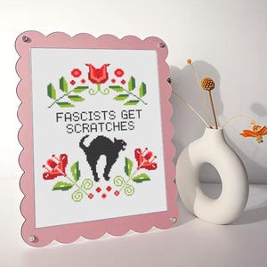 Könnte beinhalten: Ein rosa Rahmen mit gewelltem Rand zeigt eine Kreuzsticharbeit mit dem Text "Fascists Get Scratches" über einer schwarzen Katzen-Silhouette. Das Design umfasst rote Blumen und grüne Blätter. Rechts steht eine weiße Vase mit Trockenblumen.