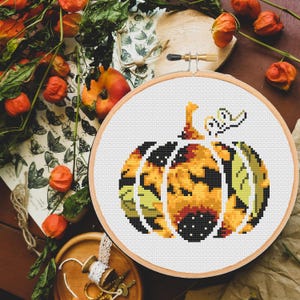 May include: A cross-stitch pattern of a pumpkin with a fall leaf design. The pumpkin is yellow, orange, and brown with black outlines. The leaves are green and brown.