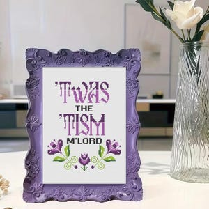 Op de afbeelding: Een ingelijst kruissteekkunstwerk met de tekst "'TWAS THE TISM M'LORD" in paars. De lijst is een decoratief, sierlijk paars met bloemendetails. Het kunstwerk heeft bloemen- en bladaccenten in groen en paars.
