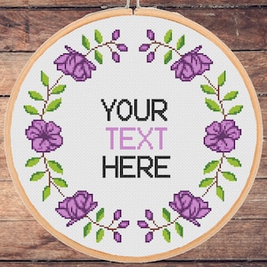 May include: A white embroidery hoop with a floral wreath design. The wreath is made of purple flowers and green leaves. The text "YOUR TEXT HERE" is in the centre of the hoop.