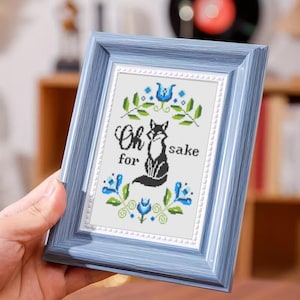 May include: A framed cross-stitch artwork with a light blue frame. The artwork features a black fox, blue floral accents, and the text "Oh for sake". The background is white, and the frame has a subtle, textured finish.