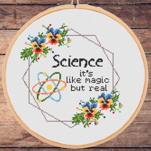 Puede incluir: Bordado de punto de cruz en un aro de madera. El diseño presenta la palabra "Science" con la frase "it's like magic but real" y un símbolo de átomo. Acentos florales en azul, amarillo y rojo rodean el texto.