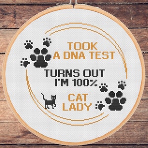 Op de afbeelding: Een witte borduurring met een goud en zwart ontwerp. Het ontwerp zegt "Took a DNA test, Turns out I'm 100% Cat Lady".