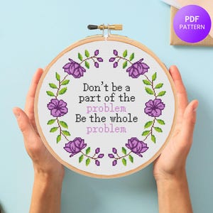 May include: A cross-stitch embroidery hoop with the text "Don't be a part of the problem, Be the whole problem." The design features purple flowers and green leaves arranged in a circular pattern. The hoop is held by hands against a light blue background.