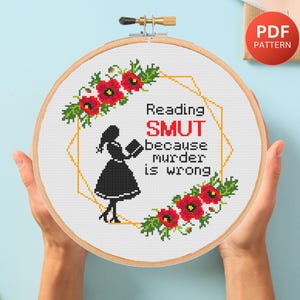 May include: A cross-stitch hoop with the text "Reading SMUT because murder is wrong." The design features a silhouette of a woman reading a book, surrounded by red poppies and green foliage. A PDF pattern is available.